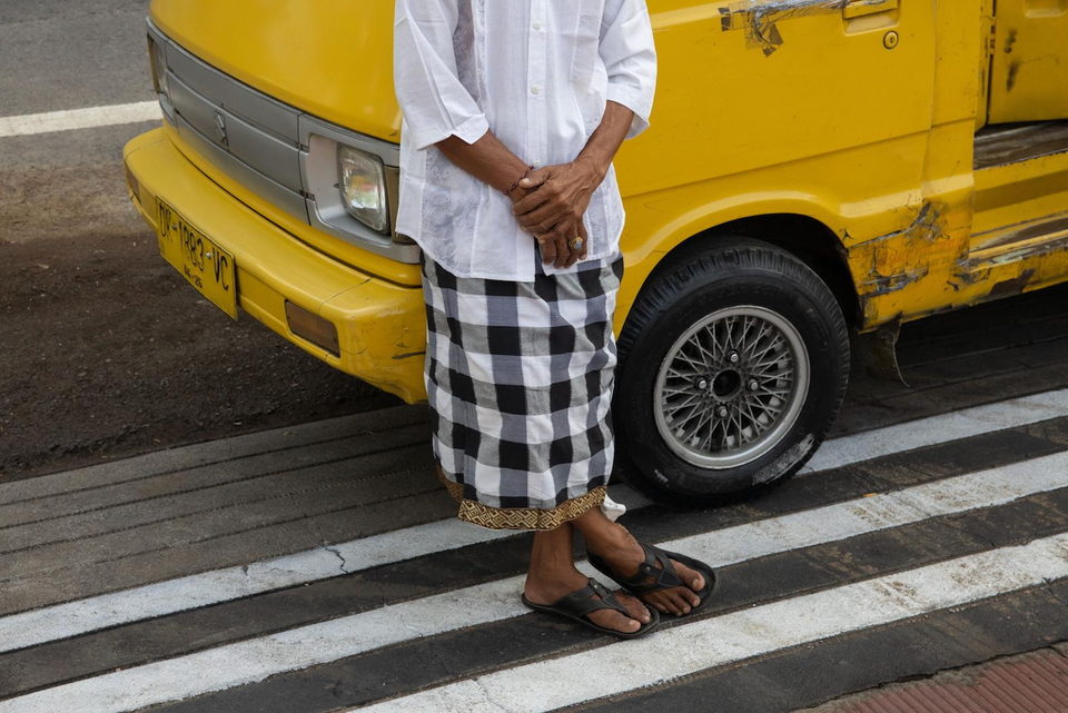 A man in a checked sarong stands at a crossing beside a yellow van.