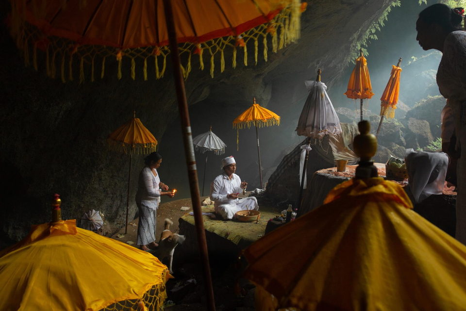 A priest sits among ceremonial umbrellas in smoky light.