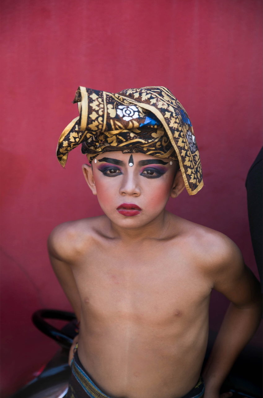 A young dancer in a gilded headdress and painted makeup against a deep pink wall.