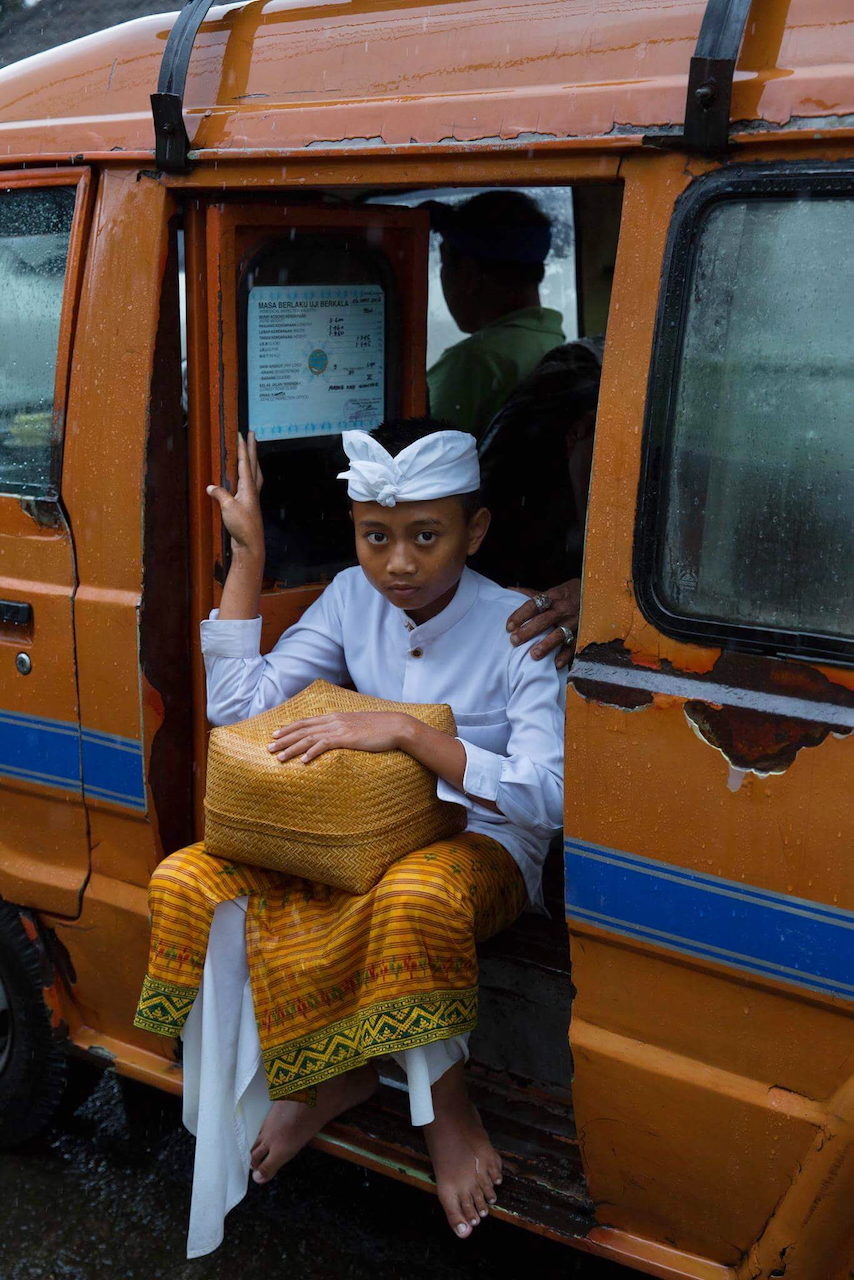 A boy in ceremonial dress sits in the doorway of an orange bus holding a hat.