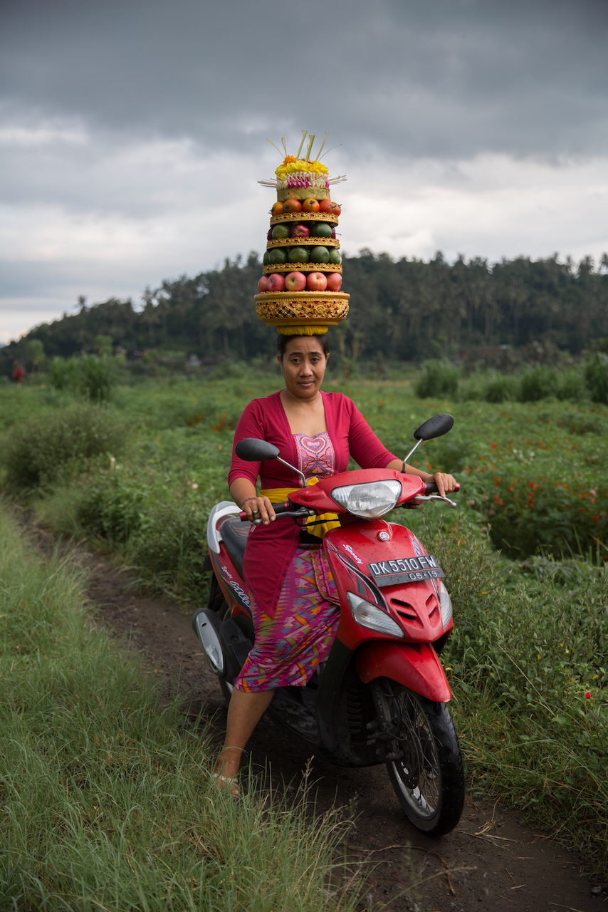 A woman on a red scooter carries a tall tiered fruit offering on her head.