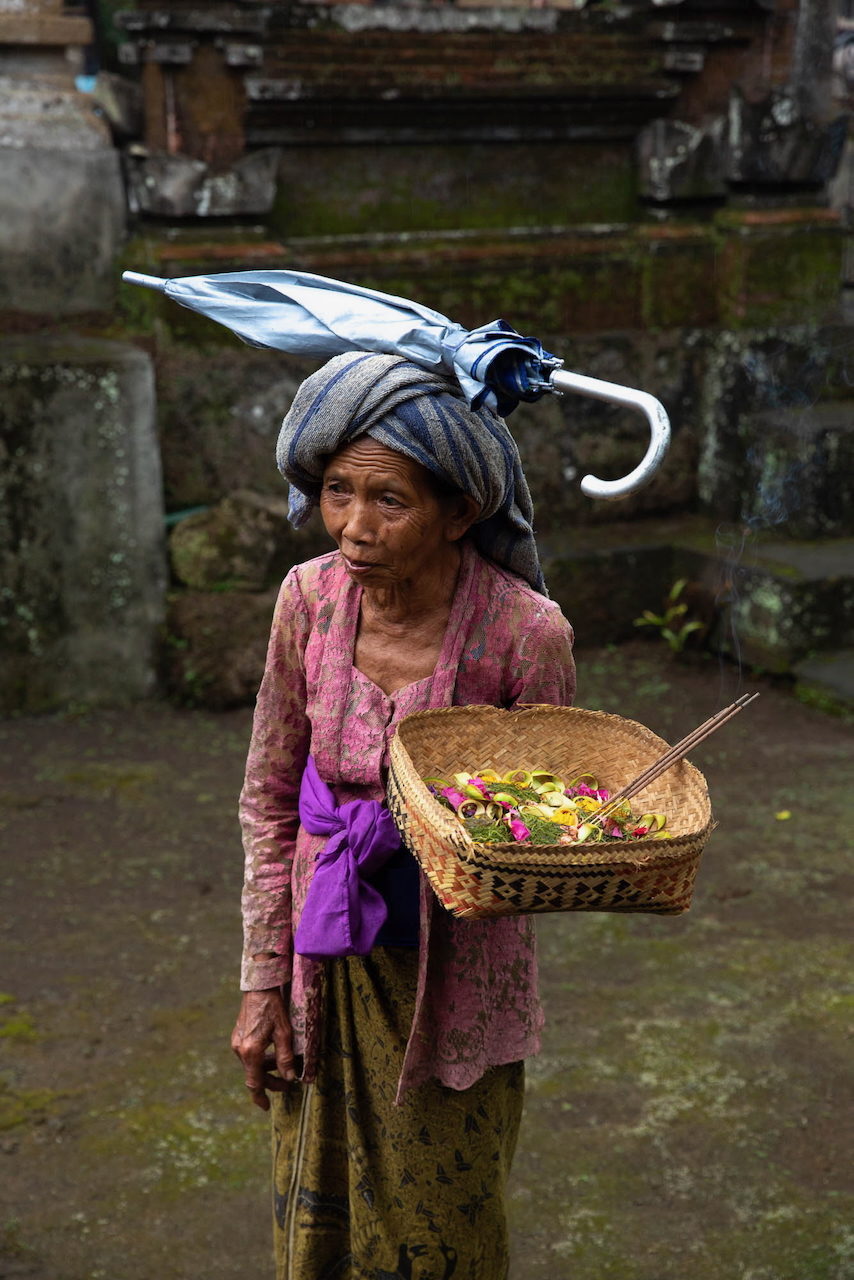 An older woman carries a basket of offerings, an umbrella folded on her head.