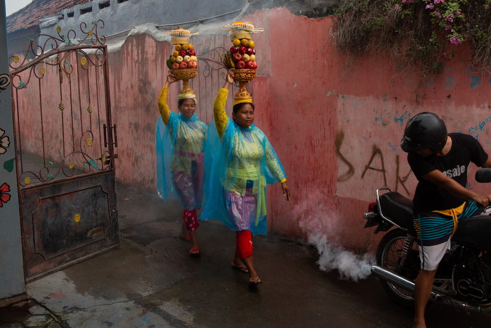Two women in blue plastic ponchos carry fruit offerings on their heads along a pink wall as a motorcyclist pulls away.
