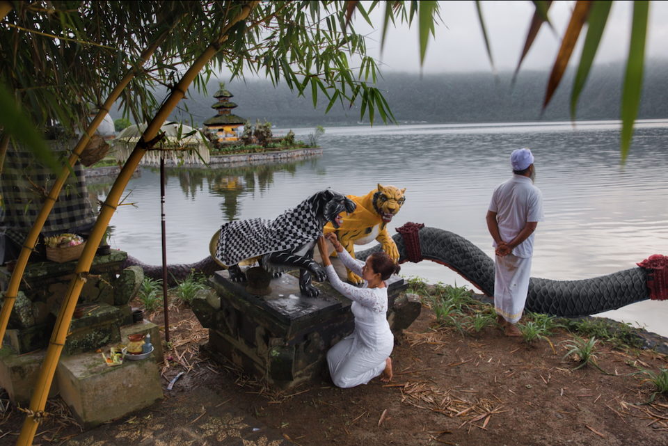 A masked barong figure is carried at a lakeside shrine.
