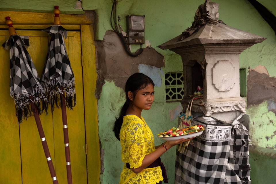 A girl in yellow carries an offering tray past a green wall and a shrine.