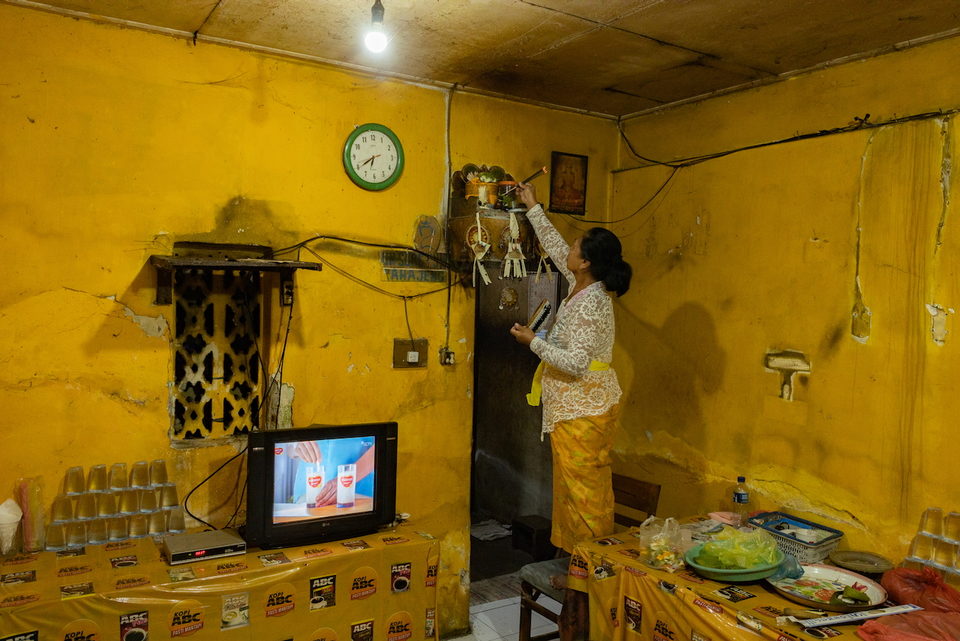 A woman reaches up to a shelf in a yellow warung, a television below.