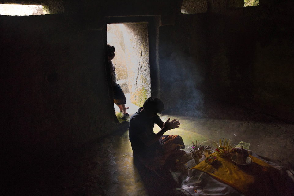 A figure kneels at offerings in a shaft of light through a stone doorway.