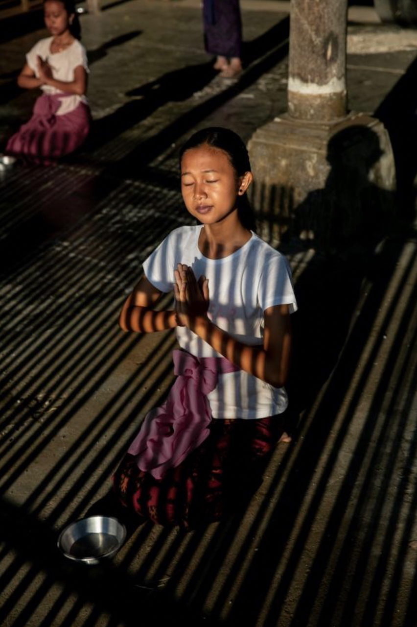 A girl prays with eyes closed, striped light across her face.