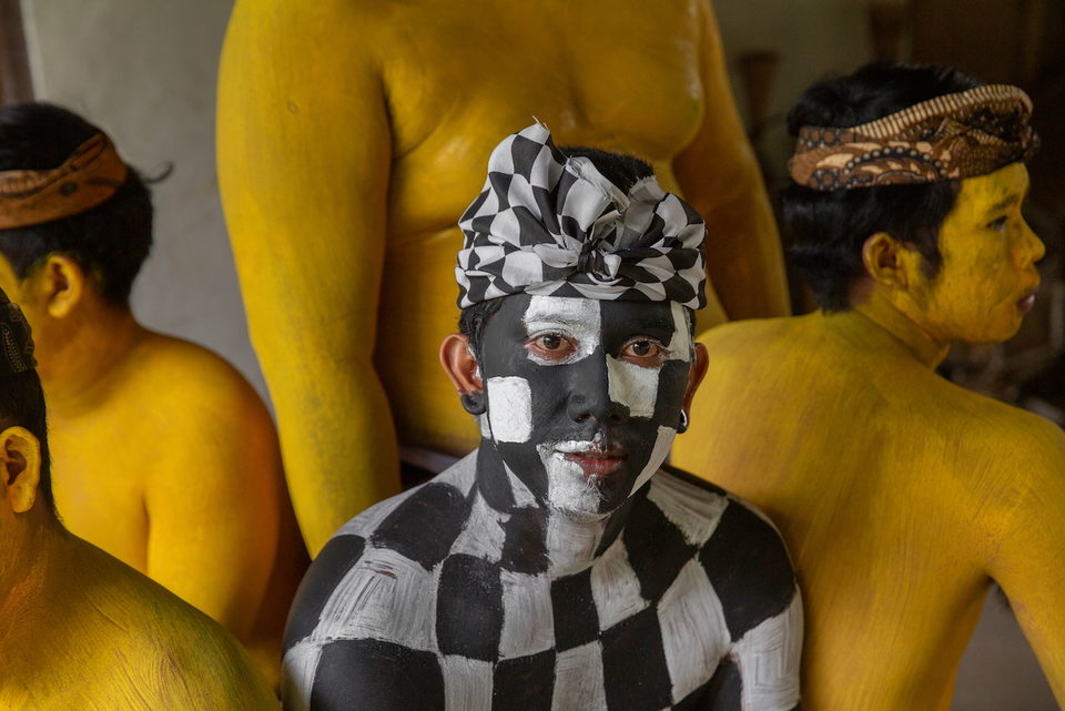 A figure painted in black and white checks stands among yellow-painted bodies.