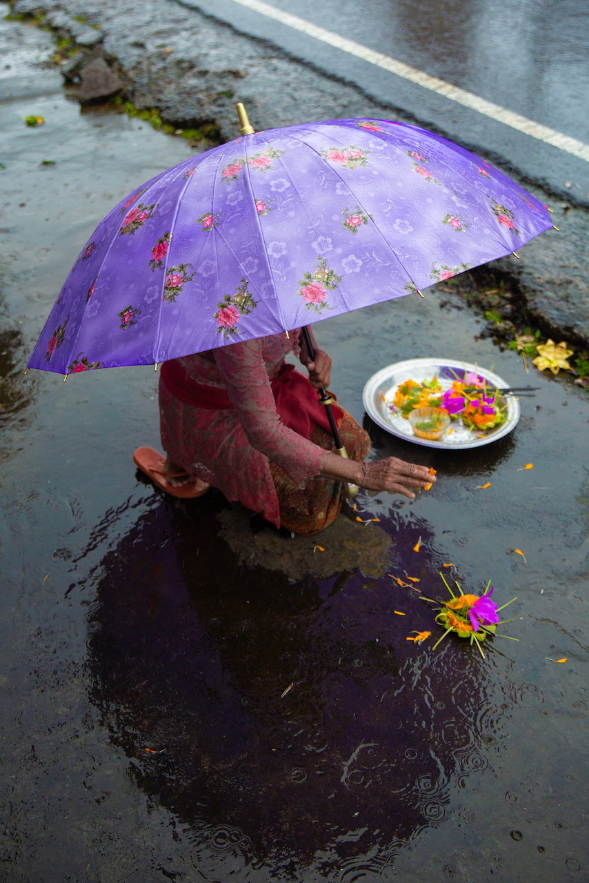 A woman places an offering into floodwater beneath a purple umbrella.