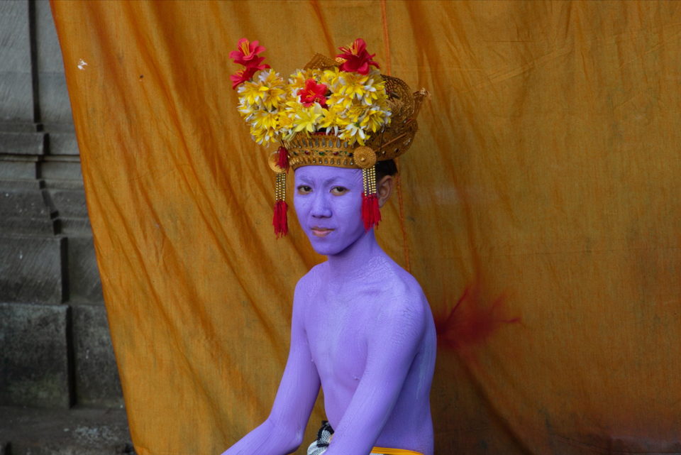 A person painted lilac in a flowered gold crown against orange cloth.
