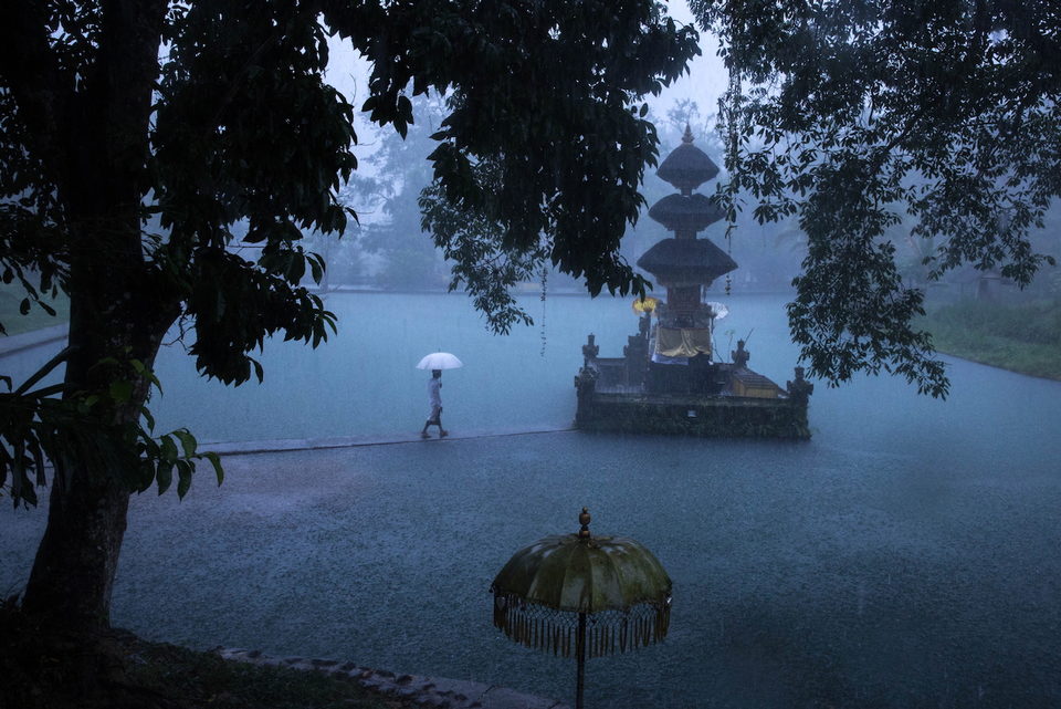 A lone figure with a white umbrella walks past a temple shrine standing in floodwater, in blue dawn mist.